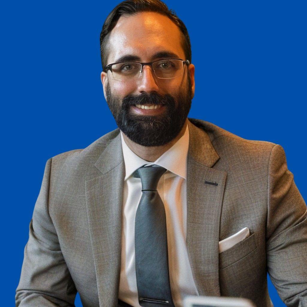 Man wearing glasses and a gray suit with a blue tie, smiling against a solid blue background.